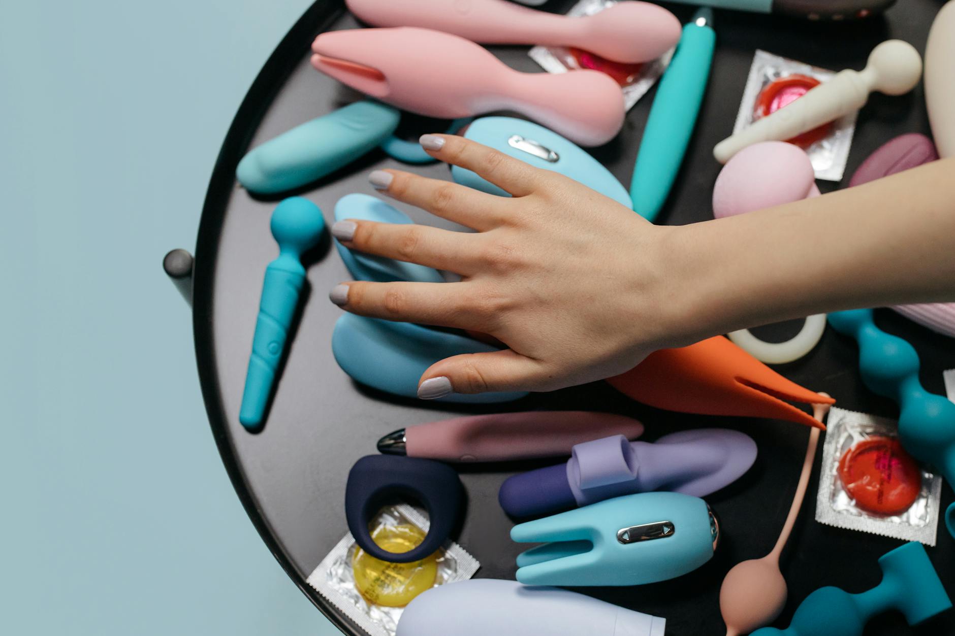 A hand reaching over a collection of colorful sex toys arranged on a table
